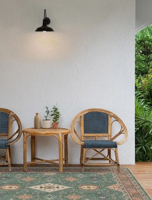 Two wicker chairs with dark cushions and a small table with potted plants are set against a white wall under a black wall lamp; greenery is visible outside.