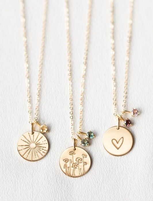 Three gold necklaces with round pendants: one with a sunburst, one with flowers, and one with a heart, each paired with a small gemstone charm, displayed on a white surface.