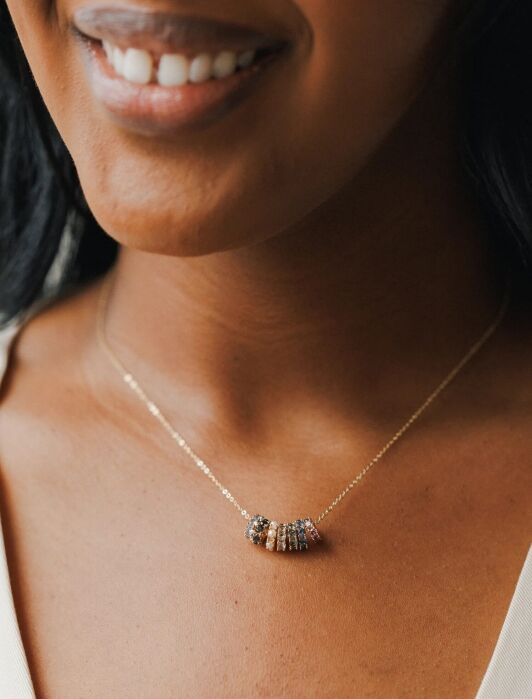 A woman wears a delicate gold chain necklace with several small, multicolored beaded charms, shown in close-up against her neck and collarbone.