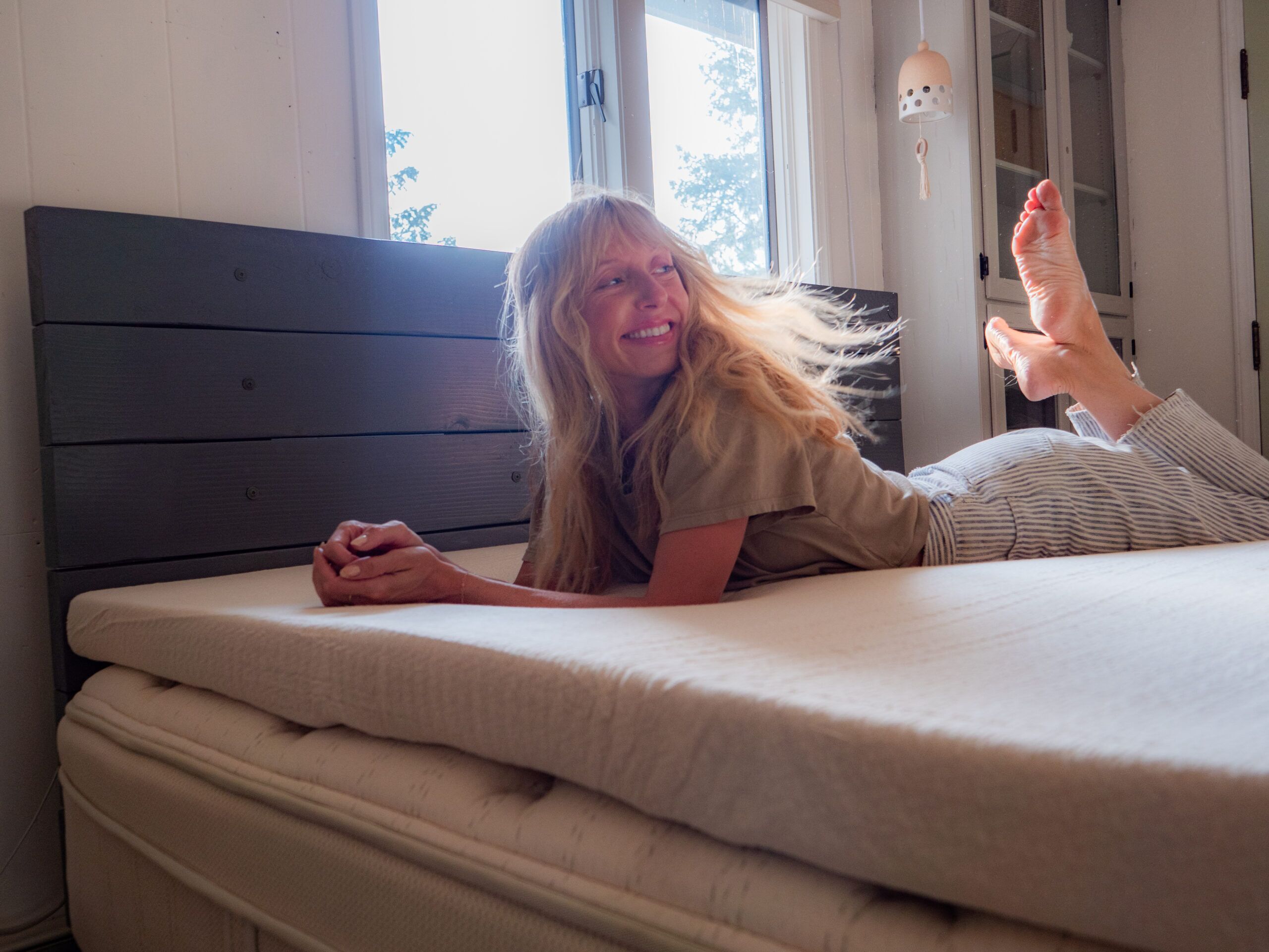 A woman with long blonde hair lies on her stomach on a bed, smiling, in a sunlit room with white walls and a window.