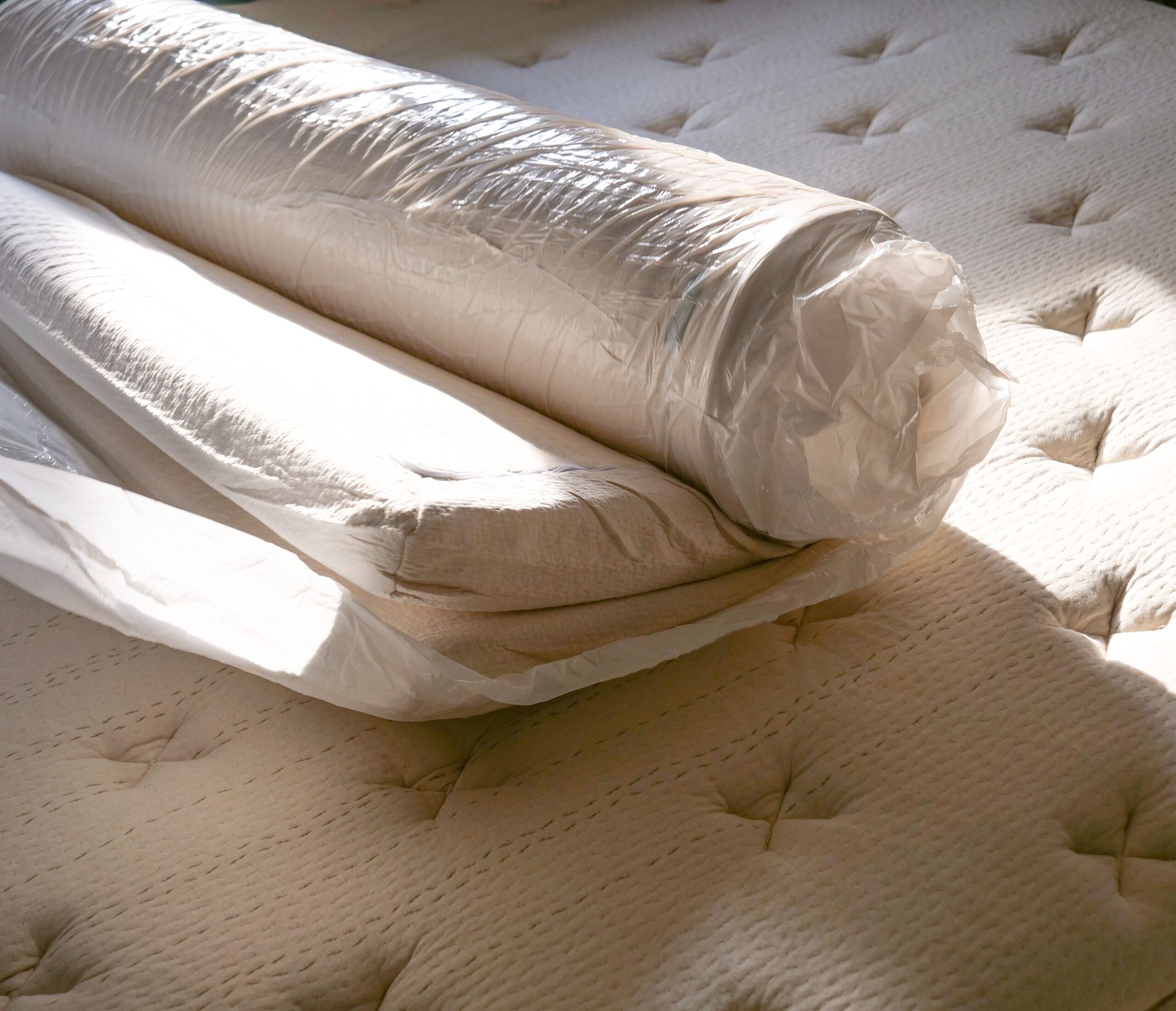 A rolled-up mattress in plastic packaging is partially unwrapped on top of an unmade mattress with a quilted surface.
