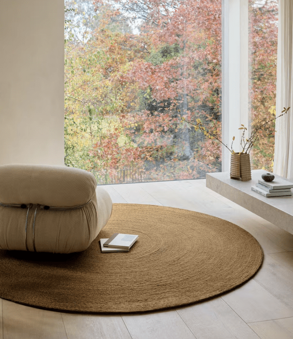 A beige lounge chair and a round woven rug are placed near a large window with a view of autumn trees. A low shelf holds a vase with branches, a bowl, and a stack of books.