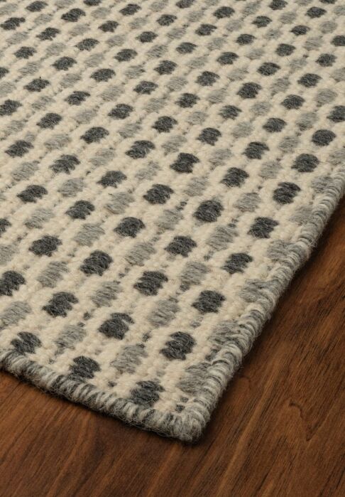 A close-up of a wool rug with a gray and cream checkered dot pattern, shown on a wooden floor.