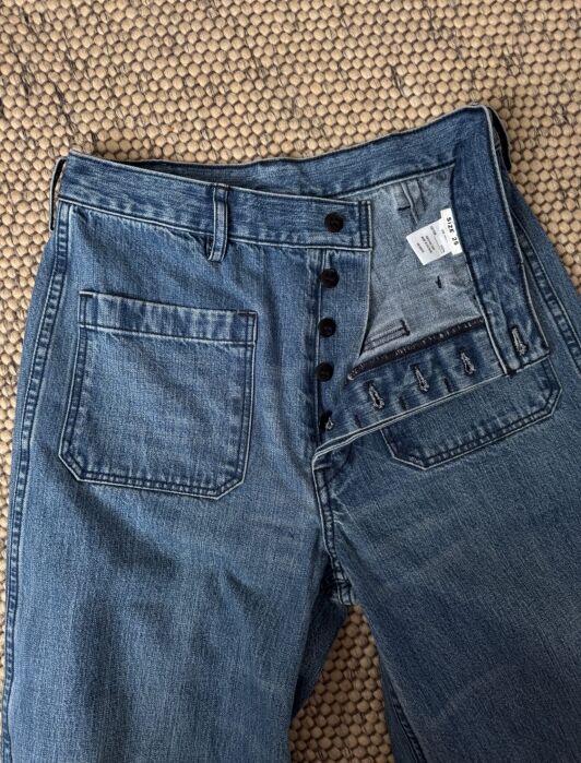 A pair of blue jeans with a button fly partially open, displayed on a textured beige surface.
