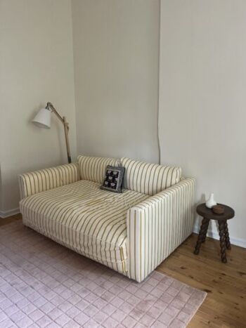 A yellow and white striped sofa with a patterned cushion sits next to a wooden side table and floor lamp on a pink rug in a room with light-colored walls and wooden floor.