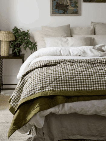 A neatly made bed with light-colored pillows and sheets, topped with a green and white checkered quilt; a side table with a plant and lamp is beside the bed.