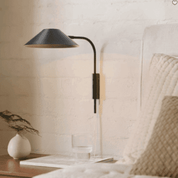 A black wall-mounted lamp is next to a bed with a beige quilt. On the wooden bedside table are a glass of water and a small vase with green foliage.