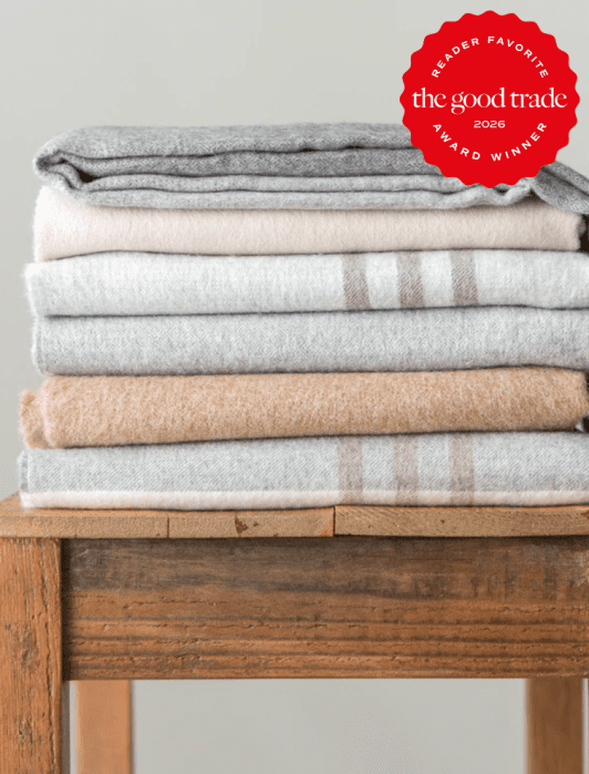A neat stack of folded neutral-colored blankets sits on a wooden stool. A red badge in the corner reads "The Good Trade Award Winner 2026 Reader Favorite.