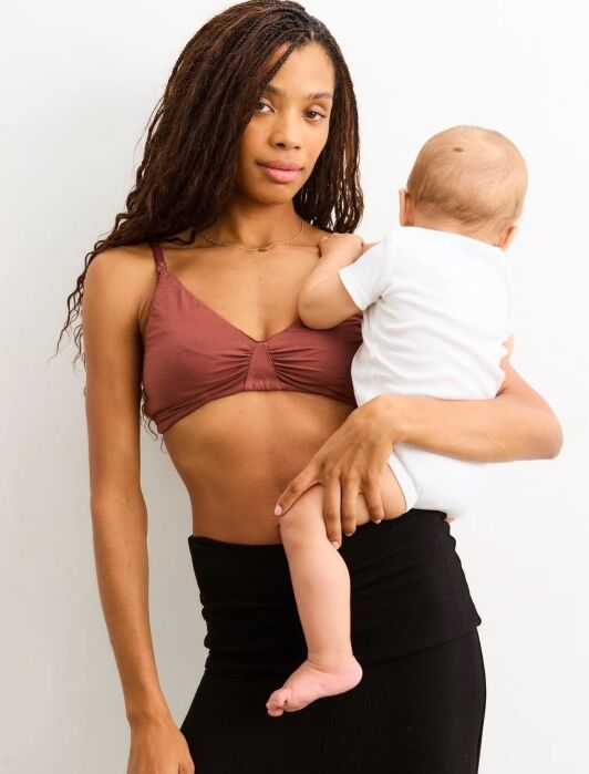 A woman in a brown bra and black skirt holds a baby dressed in a white onesie, standing against a plain white background.