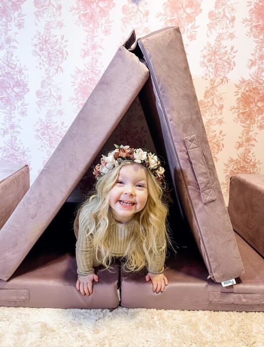 A smiling child wearing a flower crown crawls out of a pink foam play tent, with floral wallpaper and a beige carpet in the background.