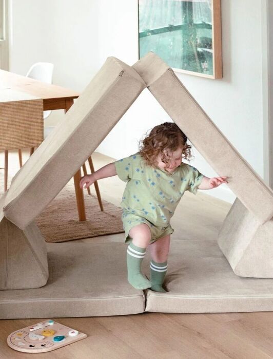 Two young children play in a living room, with one inside a tent-like structure made of foldable cushions and the other running nearby. A dining table is visible in the background.