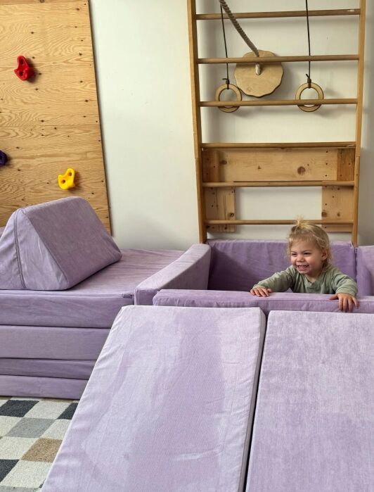 A young child smiles while playing in a soft, light purple foam playset in a playroom with a wooden climbing wall and gym equipment.