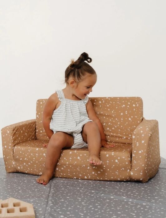 A young child sits on a small, tan, patterned sofa placed on a gray play mat, surrounded by wooden toys in a minimal indoor setting.