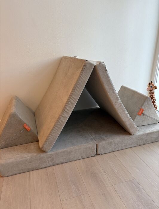 A foldable foam play couch is arranged as a tent with two triangular cushions on the sides. A toy giraffe is visible in the background near the wall.
