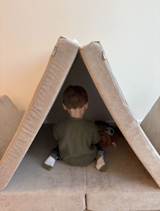 A young child sits with their back to the camera inside a makeshift tent made of beige couch cushions, playing with stuffed animals.