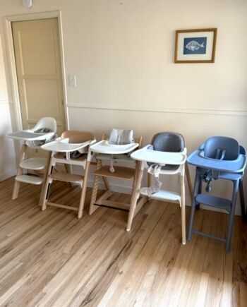 Four high chairs in different styles and colors are lined up against a wall on a wooden floor, with a framed fish print hanging above.