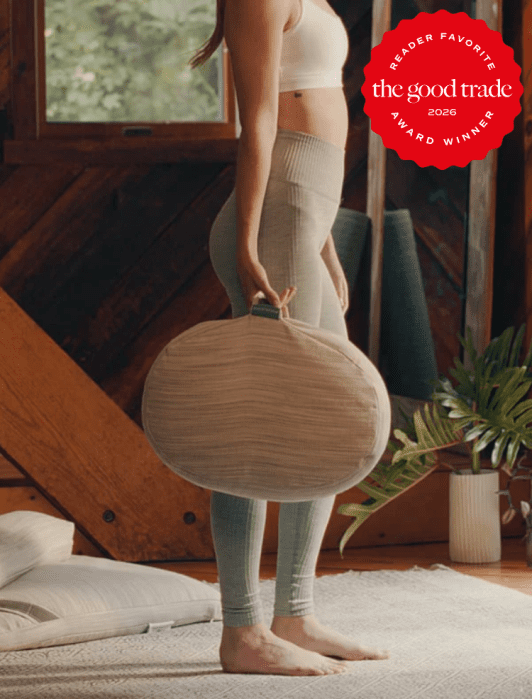 A woman in a sports bra and leggings stands indoors holding a round meditation cushion by its handle. A red award badge appears in the top right corner.