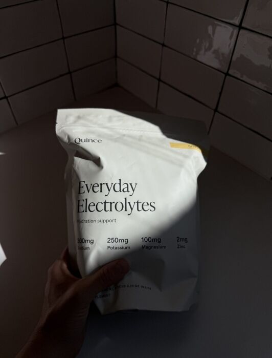 A hand holds a white bag labeled "Everyday Electrolytes" by Quince, listing sodium, potassium, magnesium, and zinc amounts. The background shows a tiled wall and counter.