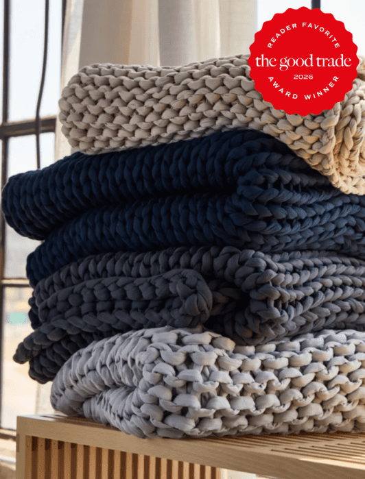 Three chunky knit blankets in cream, navy, and gray are neatly stacked on a wooden surface, with a red “The Good Trade 2026 Award Winner” badge in the top right corner.