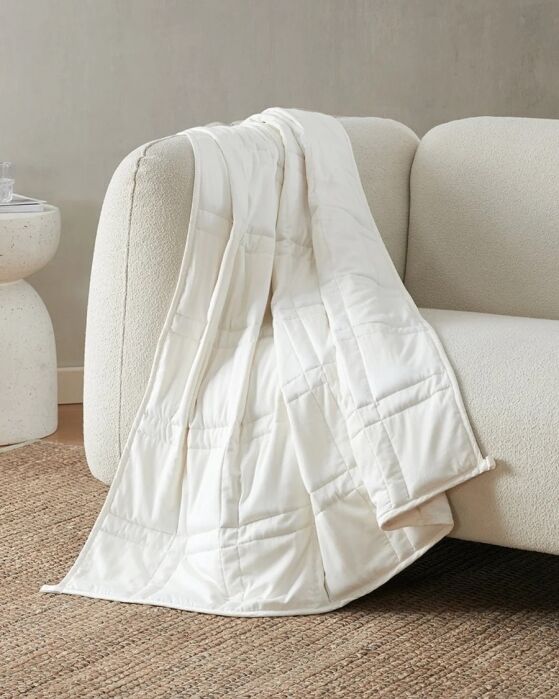 A white quilted blanket is draped over the arm of a light-colored sofa, next to a round side table, on a woven rug in a neutral-toned room.