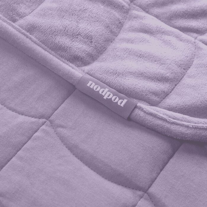 Close-up of a quilted fabric product with a visible “nodpod” label attached to the seam. The image has a purple color filter.