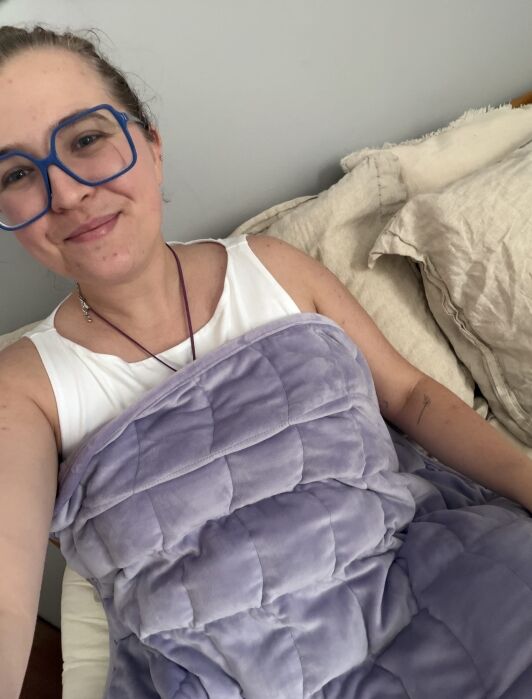 Person with blue glasses and a white top sits on a bed, smiling, covered with a light purple weighted blanket.