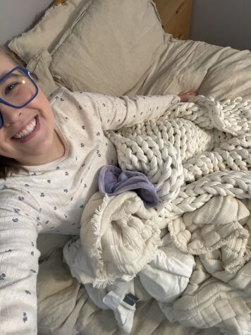 Person wearing glasses and a patterned sweater smiles while lying on a bed with cream-colored blankets and a chunky knit throw.