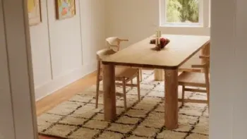A wooden dining table with three chairs sits on a patterned rug in a bright room with light walls, artwork, and a window showing greenery outside.