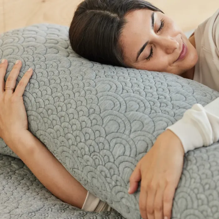A woman lies on a bed hugging a large, textured gray pillow with her eyes closed and a relaxed expression.