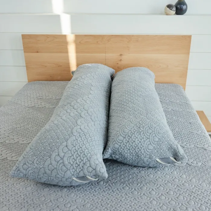 Two long, cylindrical gray pillows with textured patterns are placed side by side on a neatly made bed with a matching textured gray bedspread.