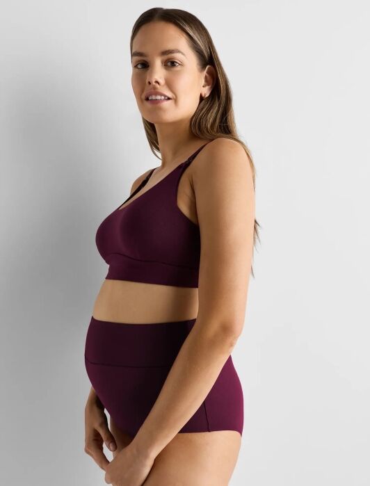 A pregnant woman in a matching burgundy bra and high-waisted underwear stands sideways, resting her hands on her belly, against a plain light gray background.