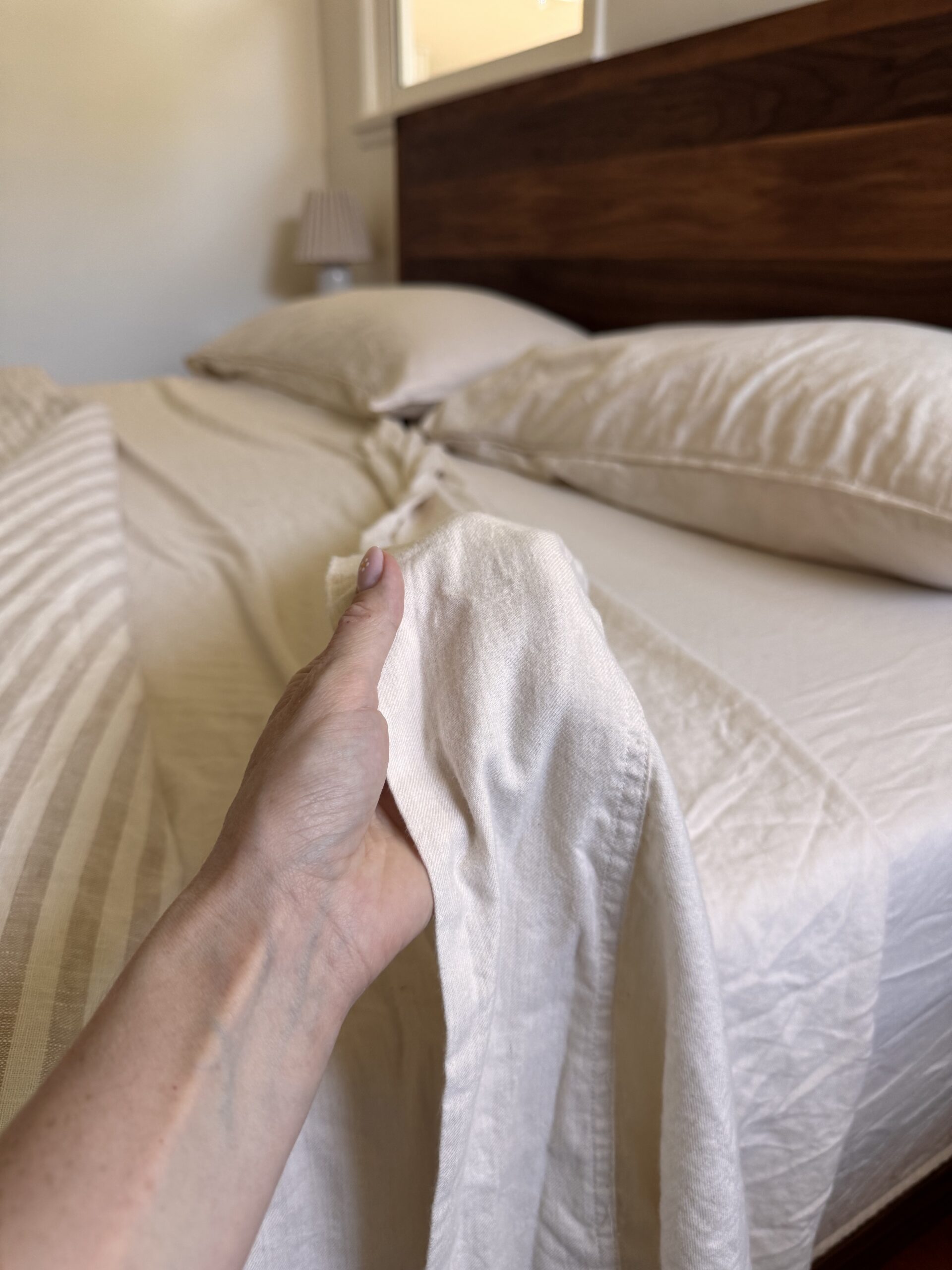 A person’s hand holds a corner of a cream-colored bedsheet while making a neatly arranged bed with matching pillows and a wooden headboard.