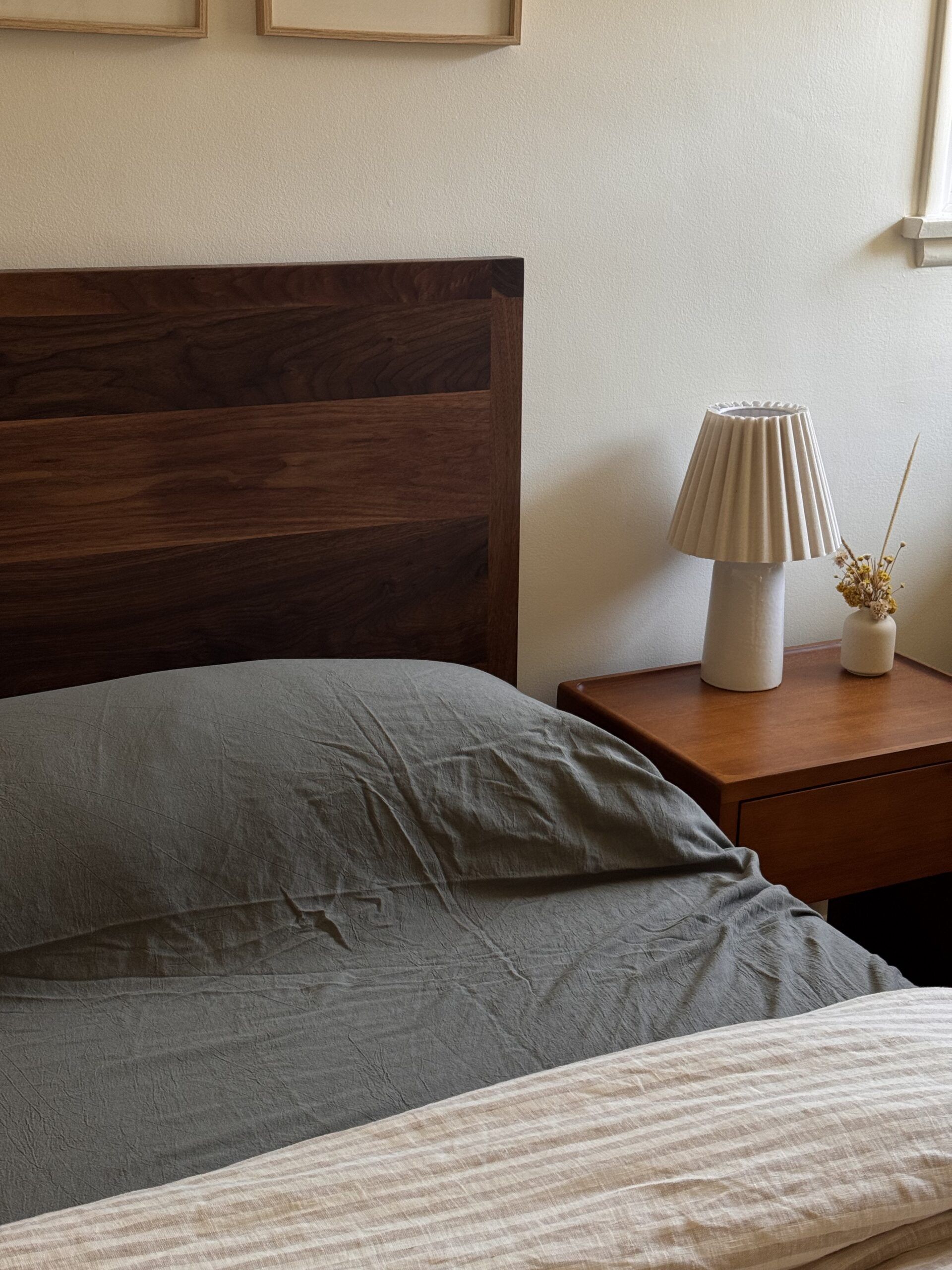 A neatly made bed with a dark wooden headboard beside a wooden nightstand holding a pleated lamp and a small vase with dried flowers.