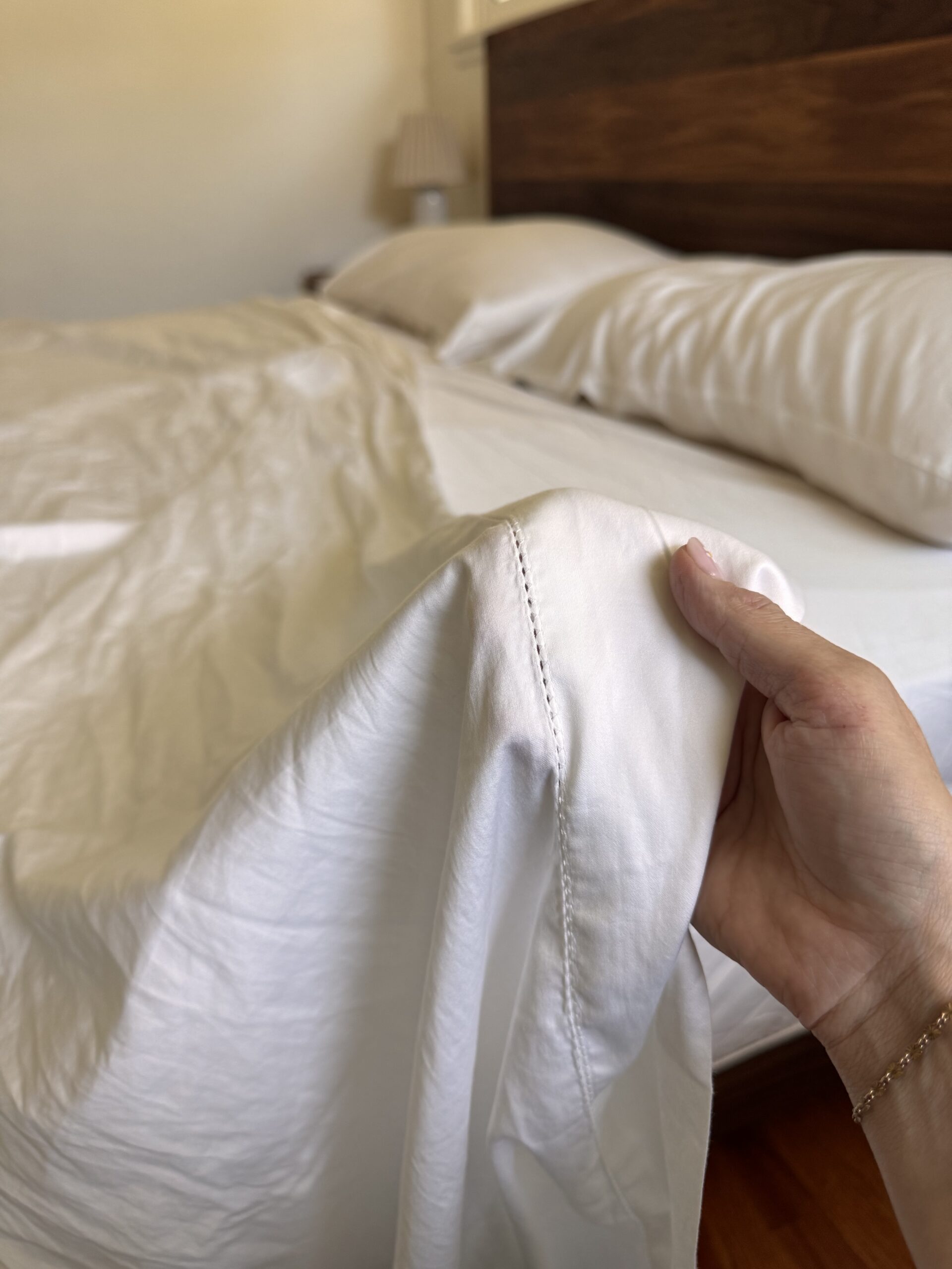 A hand holds the corner of a white bed sheet near a neatly made bed with white pillows and a wooden headboard.