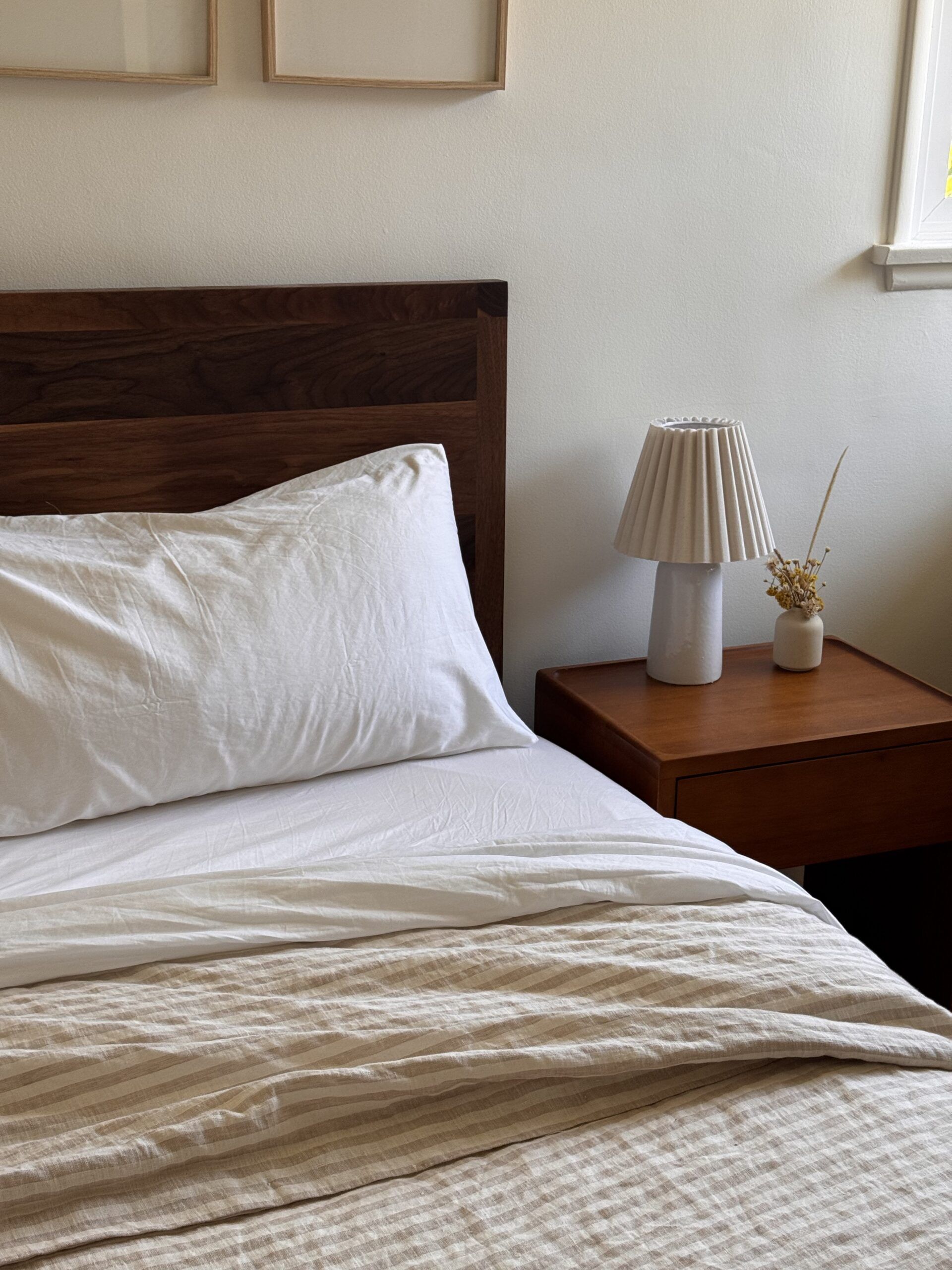 A neatly made bed with white linens next to a wooden nightstand holding a small lamp and a vase with dried flowers in a softly lit room.