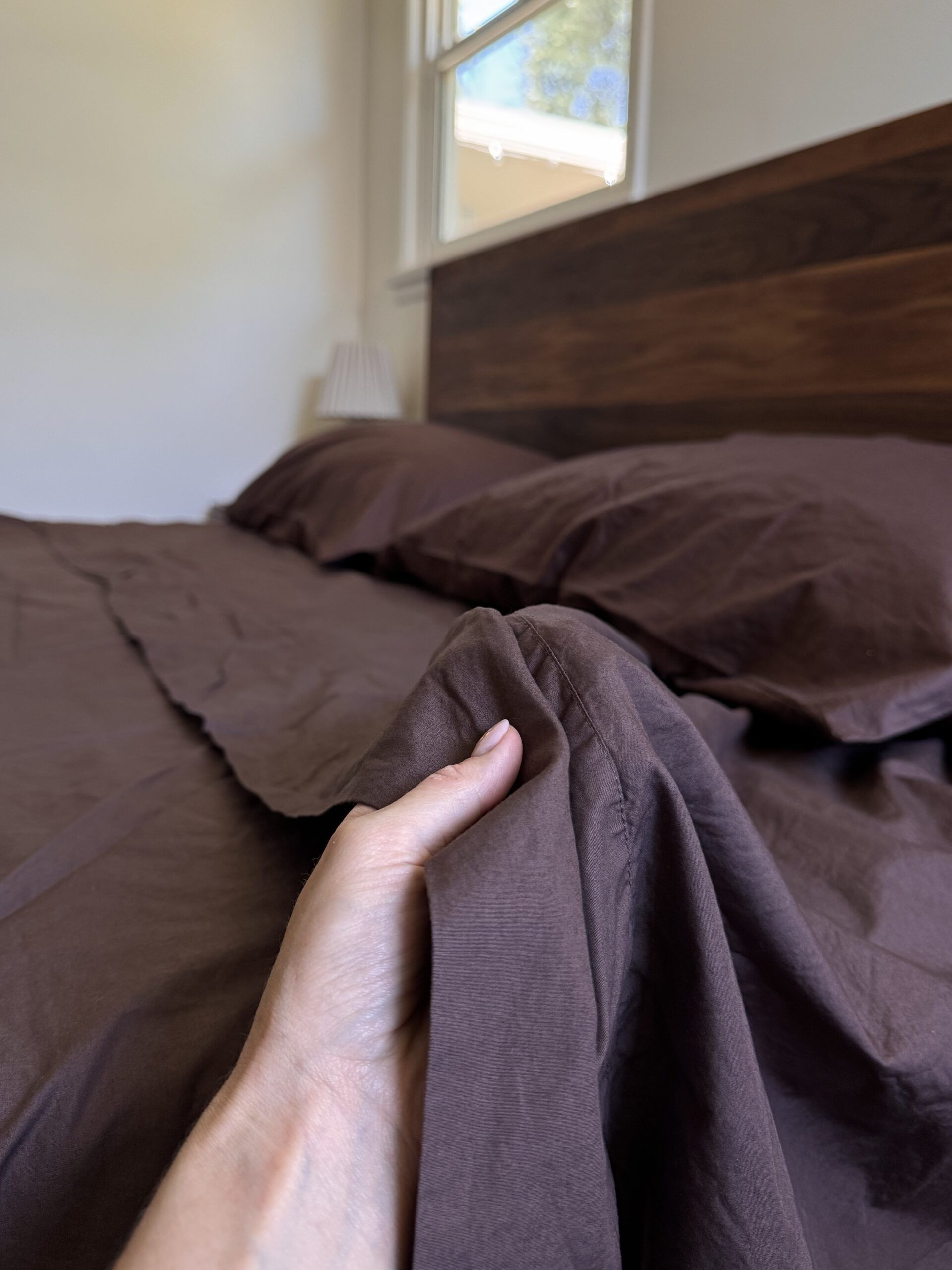 A hand holding the edge of a brown sheet on a neatly made bed with matching brown pillows, next to a wooden headboard and a window.