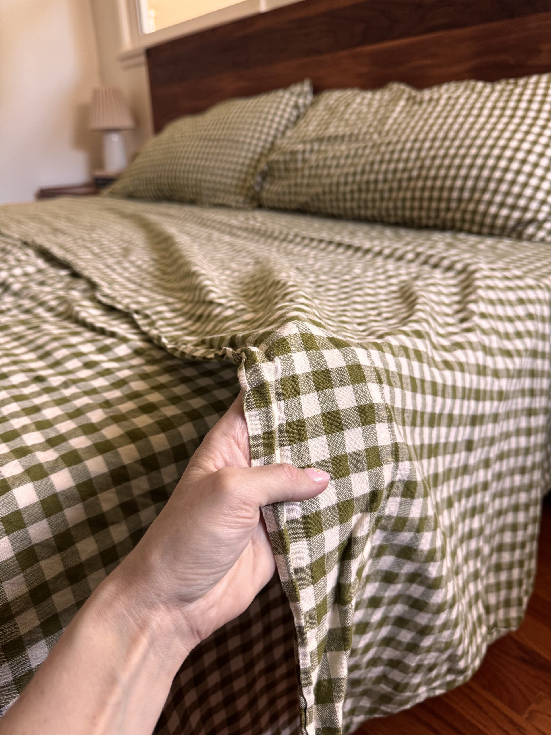 A hand holds the corner of a green and white checkered bedspread on a neatly made bed with matching pillows and a wooden headboard.