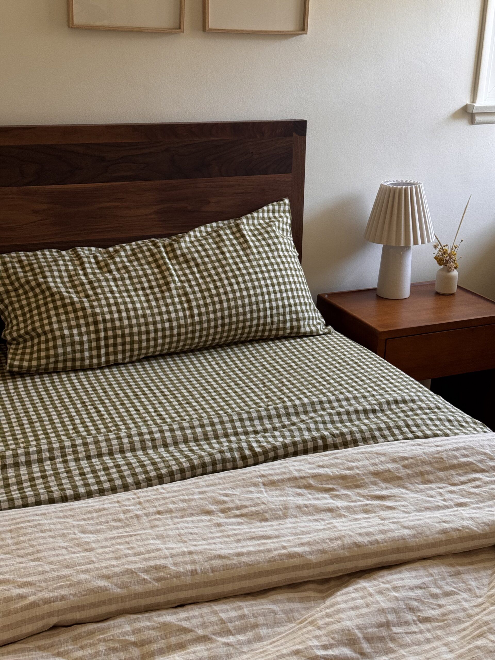 A neatly made bed with green checkered bedding, a wooden headboard, a matching wooden nightstand, a small lamp, and a dried floral arrangement.