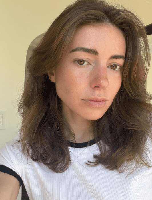 A person with long brown hair and light skin, wearing a white shirt with black trim, looks at the camera in natural lighting.