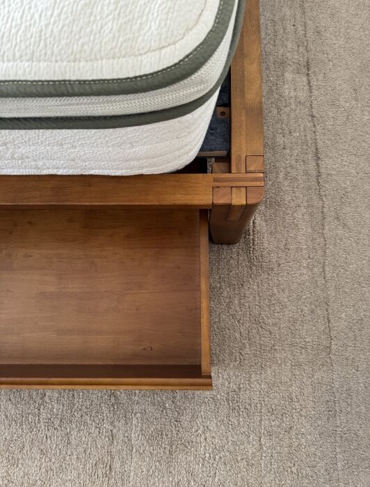 A corner of a wooden bed frame with an open storage drawer, positioned next to a mattress on a carpeted floor.