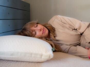A woman with long hair is lying on her side on a bed, resting her head on a white pillow and wearing a beige robe.