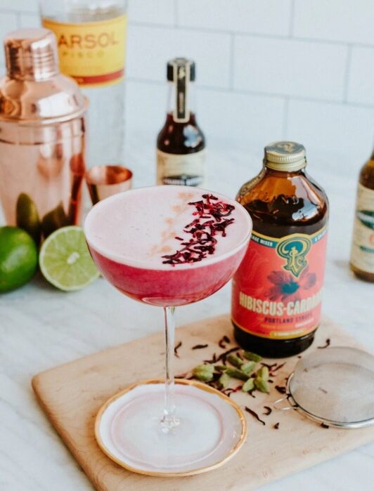 A pink cocktail in a coupe glass is garnished with dried flower petals, set on a wooden board with limes, a bottle of Hibiscus liqueur—just the look to inspire your next online shopping for bar essentials.