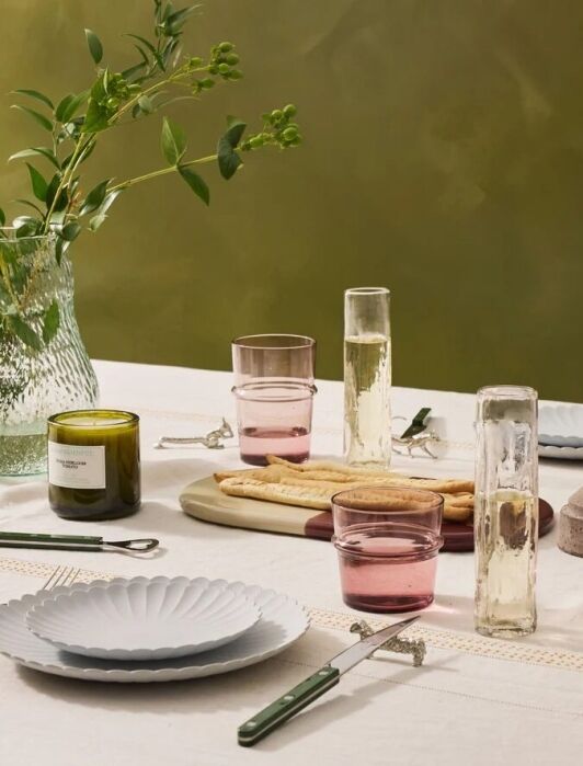 A set dining table with plates, glasses, cutlery, a green vase with foliage, a candle holder, a green glass candle, and bread on a wooden board—perfect inspiration for your next online shopping spree.