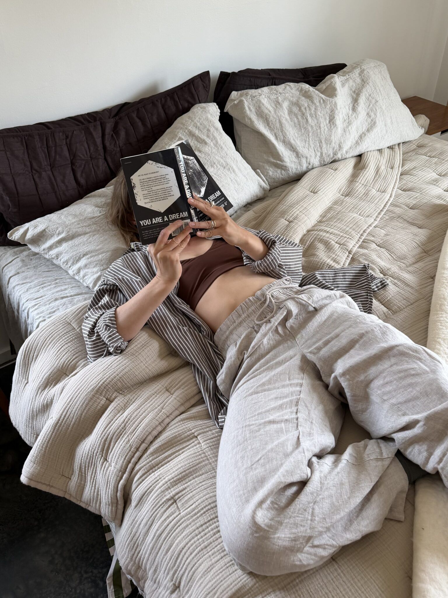 Person lying on a bed, wearing loose clothes, holding and reading a book titled "You Are a Dream" with their face partially covered by the book.