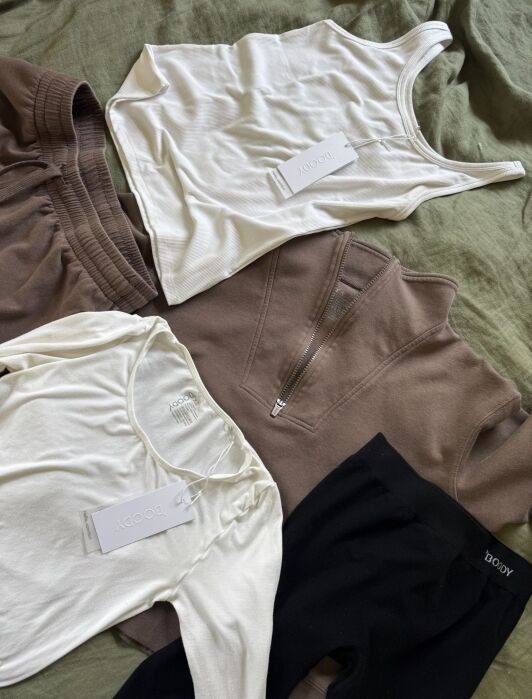 A neatly arranged flat lay of neutral-toned clothing items, including tops, shorts, and leggings, all with brand tags reading "Boody," displayed on a green fabric background.