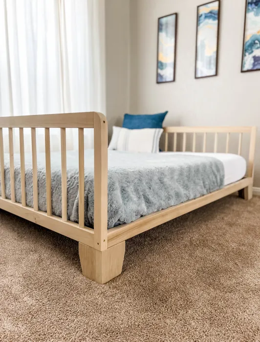 A wooden bed frame with a light gray blanket, white and blue pillows, on a beige carpet, in a room with three abstract wall art pieces and a window with sheer curtains.