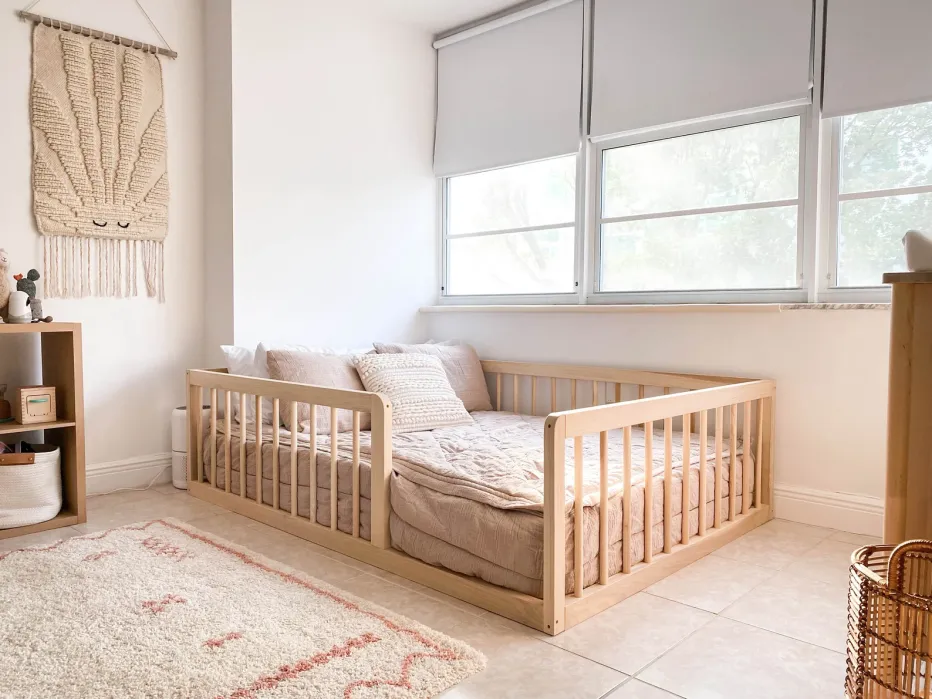 A minimalist bedroom with a low wooden bed, beige bedding, a woven wall hanging, a small shelf with toys, and large windows with roller blinds.