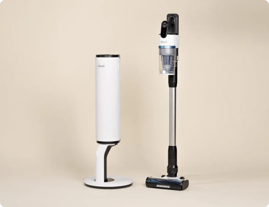 A cordless stick vacuum cleaner stands upright next to its white charging and storage base on a plain beige background.