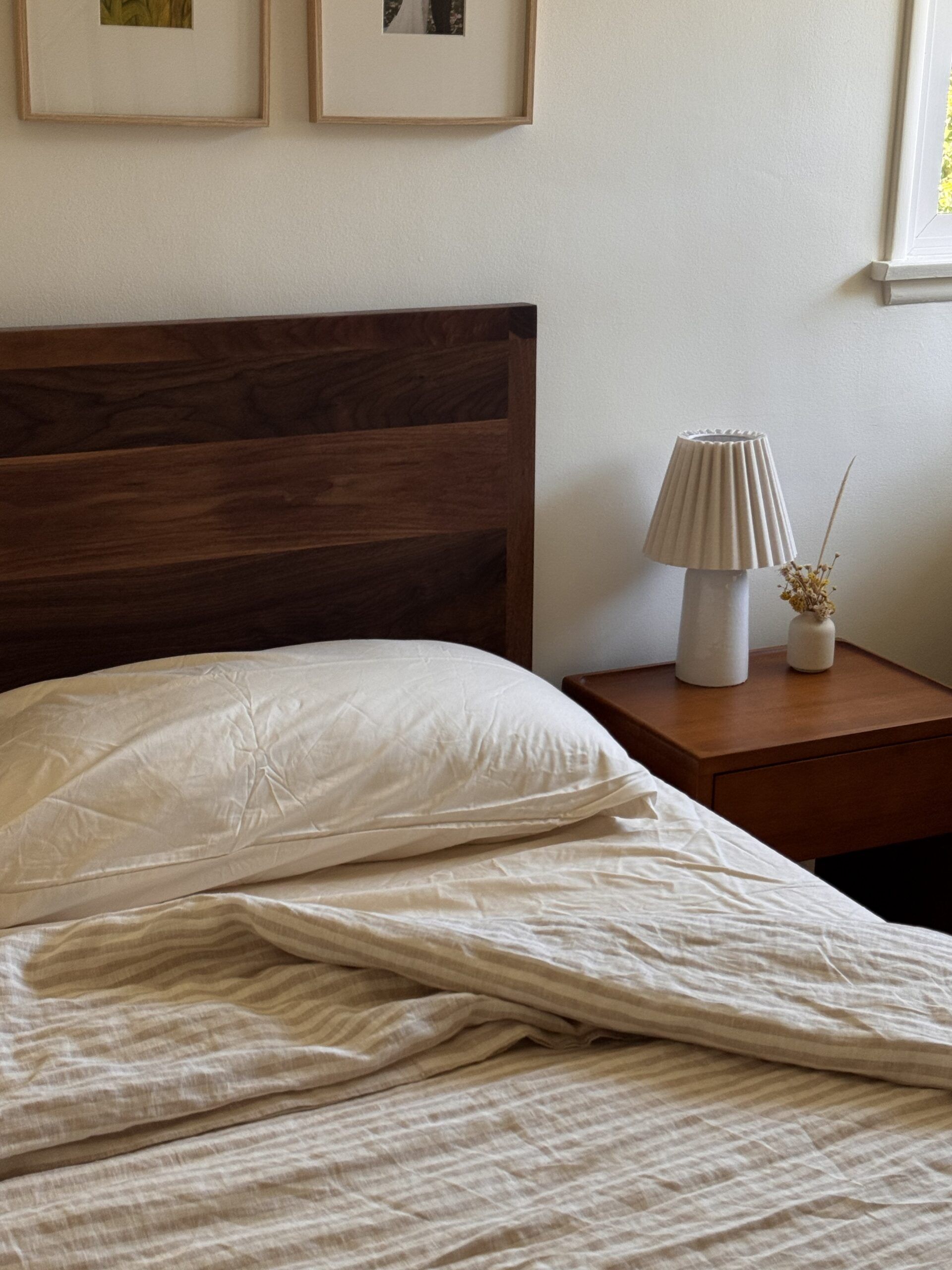 A neatly made bed with beige linens next to a wooden nightstand holding a lamp and dried flowers, with framed pictures on the wall above.