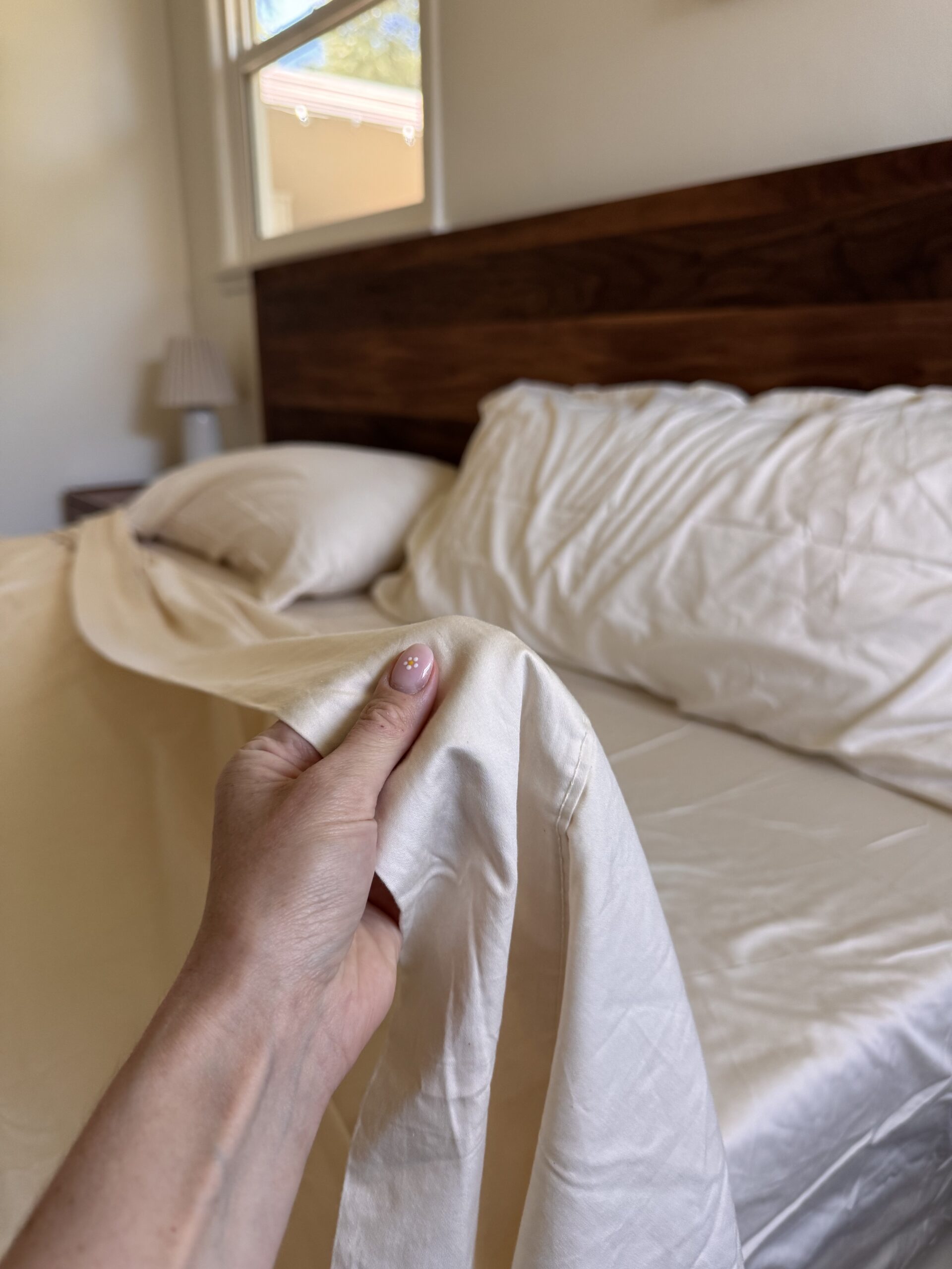 A person’s hand holds a corner of a cream-colored bedsheet near a neatly made bed with matching pillows and a wooden headboard.